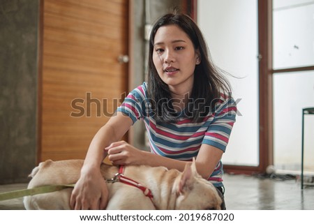 Pet lover, an Asian casual woman sits comfortably on the house floor, hugging a young french bulldog by hand. This cute tame dog had a happy relationship with her friend and family.