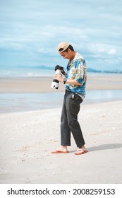 Petchaburi,Thailand -July 10,2021 :Candid Of Young Attractive Asian Man Hold Camera Taking Photo In Sea View Background. Happy Asian Hipster Male Photographer In Youth Freedom Culture Lifestyle Travel