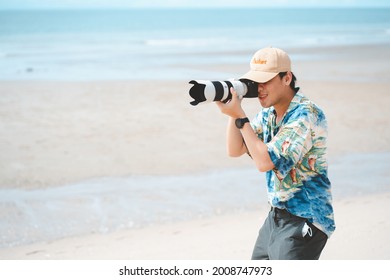 Petchaburi,Thailand -July 10,2021 :Candid Of Young Attractive Asian Man Hold Camera Taking Photo In Sea View Background. Happy Asian Hipster Male Photographer In Youth Freedom Culture Lifestyle Travel