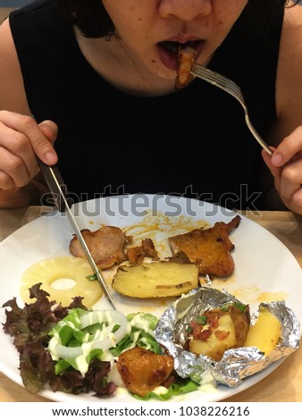A petite Asian woman wearing a black blouse is eating spicy chicken steak in a white round plate served with fresh vegetables and buttered potato.
