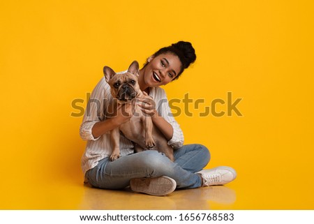 Pets And People. Young Beautiful Afro Woman Posing With Ger Cute Dog, Sitting On Fllor In Studio Over Yellow Background, Copy Space