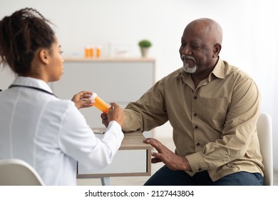 Pharmaceutical Drug Prescription Concept. Young Black Doctor Showing Elderly Male Patient Pills Bottle, Pointing At Tablets. Female Specialist Prescribing Mature Man Treatment, Painkiller Or Vitamin