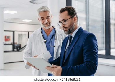 Pharmaceutical sales representative talking with doctor in medical building, presenting new medication on tablet. Hospital director, manager in modern clinic with surgeon.