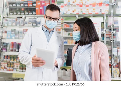 Pharmacist In Eyeglasses And Medical Mask Holding Digital Tablet Near Asian Woman In Drugstore