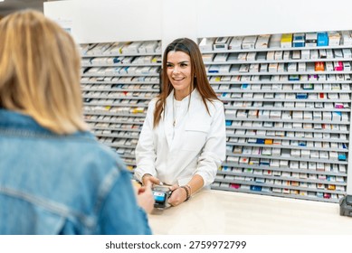 Pharmacist smiling while assisting customer with a contactless card payment at pharmacy counter, shelves of medications visible, representing modern retail healthcare service