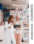 Pharmacist is using a digital tablet to show information about a medication to a customer in a pharmacy. The pharmacist is wearing a white coat and the customer is dressed casually