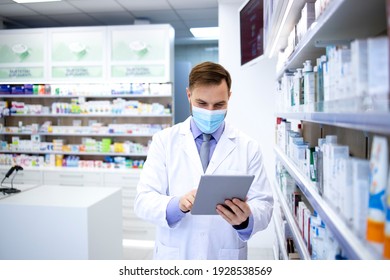 Pharmacist Wearing Face Mask And White Coat Working In Pharmacy Store On Tablet Computer During Corona Virus Pandemic. Healthcare And Medicine.