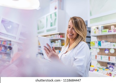 Pharmacist working with a tablet computer in the pharmacy holding it in her hand while reading information. Cheerful happy pharmacist chemist woman working in pharmacy drugstore with tablet computer - Powered by Shutterstock