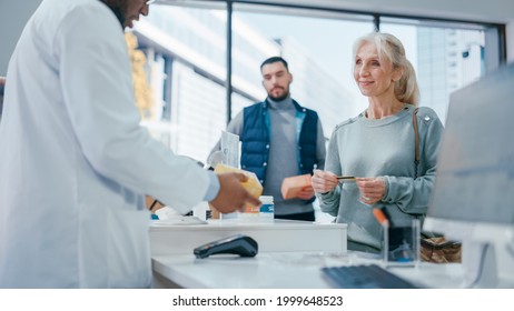 Pharmacy Drugstore Checkout Cashier Counter: Black Male Pharmacist Service Senior Female Customer Buying Prescription Medicine Package And Paying Using Contactless Credit Card Holding It To Terminal