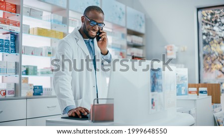 Pharmacy Drugstore Counter: Portrait of Helpful Black Male Pharmacist Talking on the Mobile Phone with Customer, Giving Medicine Recommendation, Uses Computer, Checks Prescription Drugs Availability