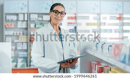 Pharmacy: Portrait of Professional Beautiful Caucasian Female Pharmacist Uses Digital Tablet Computer, Checks Medicine Inventory, Looks at Camera, Smiles Charmingly. Drugstore Health Care Products