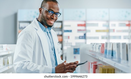 Pharmacy: Portrait Of Professional Confident Black Pharmacist Uses Digital Tablet Computer, Checks Inventory Of Medicine, Looks At Camera And Smiles Charmingly. Drugstore With Health Care Products