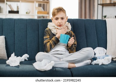 Pharyngitis, Sore Throat Concept. Sick Caucasian Teenage Boy Touching Sore Throat, Feeling Unwell, While Sitting On The Sofa At Home, And Drinking Hot Tea.