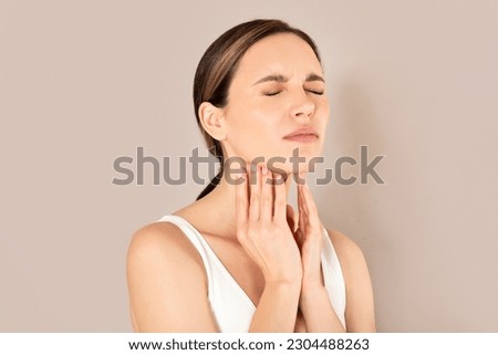 Pharyngitis, tonsillitis. Sad young european woman in white top has painful throat when swallowing, touching her neck, isolated on beige studio background, panorama with copy space