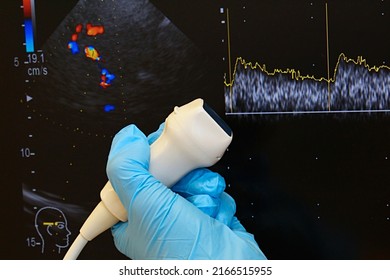 Phased array ultrasound diagnostic probe held in left hand of doctor in blue medical glove, transcranial neurological doppler examination with curves on USG display in background