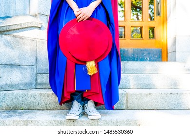 PhD (doctoral) Graduate In Regalia Gown, Holding Tudor Bonnet Cap, Sitting On University Steps, With Sneaker Canvas Shoes Showing. Red And Blue Grad Gown, Gold Tassle Showing.