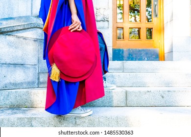 PhD (doctoral) Graduate In Regalia Gown, Holding Tudor Bonnet Cap, Sitting On University Steps, With Sneaker Canvas Shoes Showing. Red And Blue Grad Gown, Gold Tassel Showing.