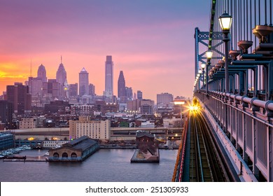 Philadelphia under a hazy purple sunset. An incoming train crosses Ben Franklin Bridge.