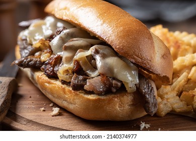 Philly Cheesesteak Sandwich Made With Steak, Cheese And Onions On A Toasted Hoagie Roll With French Fries On A Wooden Board