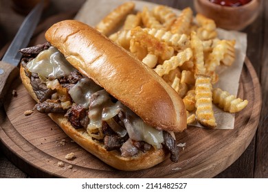 Philly Cheesesteak Sandwich Made With Steak, Cheese And Onions On A Hoagie Roll With French Fries On A Wooden Board