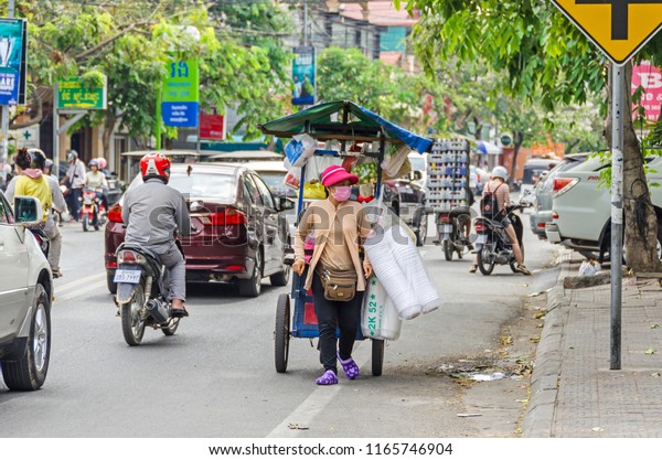 Phnom Penh, Cambodia - April\
8, 2018: View of the typical Cambodian street with many trees,\
mopeds, cars and a female street vendor with her mobile sales\
stand