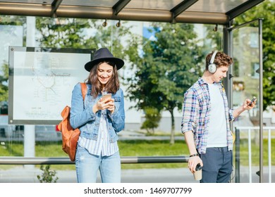 Phone Addict Youth, Modern Lifestyle Concept. One Of Teenager Couple Listening To Music With Headphone, Other Browsing Internet With Smartphone In Tram Stop Outdoor No Attention To One Another.