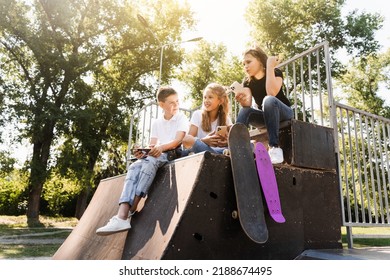 Phone Addicted Sports Children With Skateboard And Penny Boards Smiling And Looking At Smartphones On Sports Ramp On Playground. Kids Addiction Of Phones