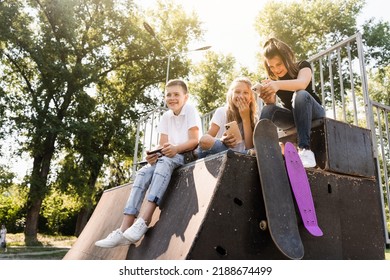 Phone Addicted Sports Children With Skateboard And Penny Boards Smiling And Looking At Smartphones On Sports Ramp On Playground. Kids Addiction Of Phones