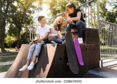 Phone Addicted Sports Children With Skateboard And Penny Boards Smiling And Looking At Smartphones On Sports Ramp On Playground. Kids Addiction Of Phones