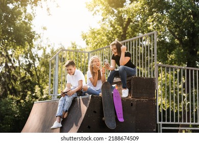 Phone Addicted Sports Children With Skateboard And Penny Boards Are Sitting And Looking At Smartphones On Sports Ramp On Playground. Kids Addiction Of Phones