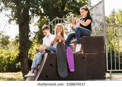 Phone Addicted Sports Children With Skateboard And Penny Boards Are Sitting And Looking At Smartphones On Sports Ramp On Playground. Kids Addiction Of Phones