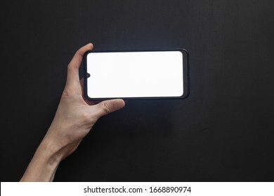 Phone with a bright screen in a horizontal position holds his hand against a dark background.