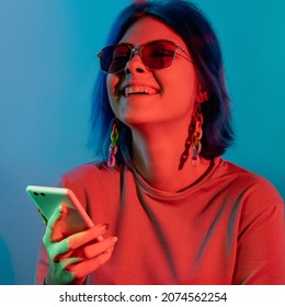 Phone entertainment. Millennial lifestyle. Mobile technology. Joyful laughing woman using smartphone in red neon light isolated on blue background.