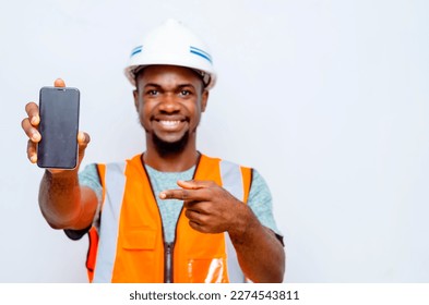 "phone in focus": young happy black male construction worker holding up mobile phone display at camera while pointing one finger at the phone - Shutterstock ID 2274543811