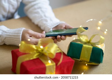 A Phone With A Green Screen In The Hands Of A Woman In A White Sweater, A Red And Green Gift Box And Bokeh Lights On The Table. Close Up. Concept Of Online Shopping For Christmas And New Year.