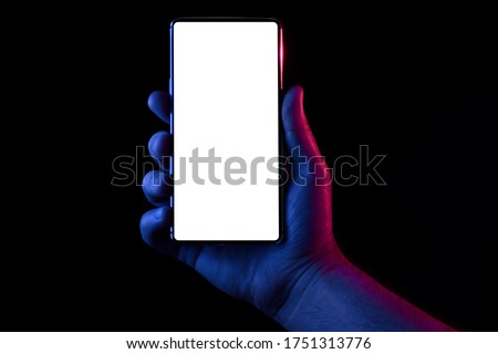 Phone in hand. Silhouette of male hand lit with blue and red neon lights holding bezel-less smartphone on black background. Screen is cut with clipping path.