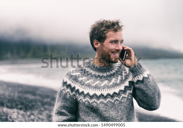 アイスランドの黒い砂浜を歩く冬のセーターでスマートフォンで話している電話男性 アイスランドの羊毛の服 テクノロジーを備えた携帯電話 の写真素材 今すぐ編集