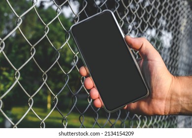 The Phone Is In The Man's Hand. Against The Background Of A Steel Wire Fence And Nature