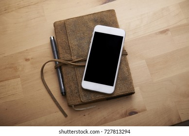 Phone On Top Of A Notebook On A Table With The Corner Of The Phone Hanging Off The Edge Of The Book Ever So Slightly. 