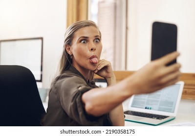 Phone Selfie With A Playful Business Woman Having Fun, Being Goofy And Joking While Working In Her Office. Young Female Sticking Out Her Tongue And Making A Face At Work While Feeling Carefree