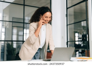 Phone talk. Busy pretty brazilian or hispanic curly stylish business woman, secretary, manager, stand near a work desk in modern office, working in a laptop and having smart phone conversation, smiles - Powered by Shutterstock