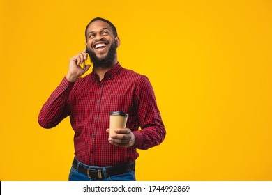 Phone Talk. Laughing black man talking on cell phone standing over yellow studio wall, holding paper coffee cup