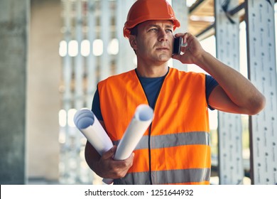 Phone Talk. Thoughtful Attentive Builder In Orange Uniform Standing With The Drawings And Having A Phone Talk