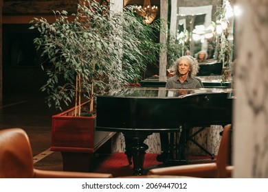 Photo of 70-year-old man plays grand piano in lobby of hotel view from afar, gray curly hair. 