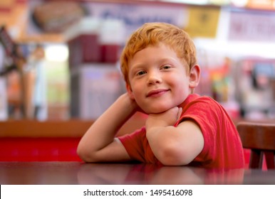 Photo Of Adorable  Little Boy With Ginger Hair. Happy Child Smiling At The Camera As He Sits At A Table Resting His Chin On His Folded Arms. Cafe, Restaurant