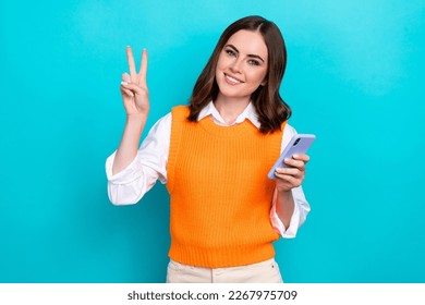 Photo of adorable positive cheerful woman bob hairstyle orange waistcoat hold phone showing v-sign isolated on teal color background