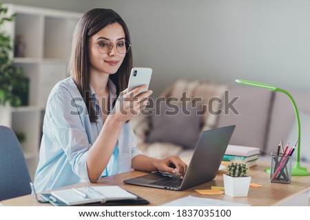 Photo of adorable young lady hold telephone look screen palm keyboard wear glasses shirt in home office indoors
