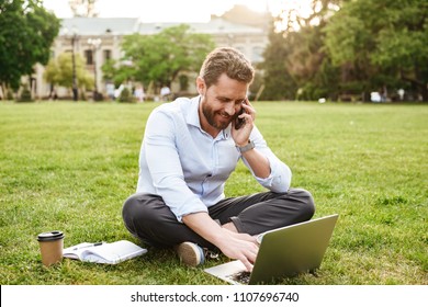 Photo Of Adult European Man In Business Clothing Sitting On Grass In Park With Legs Crossed And Speaking On Mobile Phone While Working On Silver Laptop