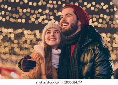 Photo of affectionate boyfriend hug girlfriend look enjoy x-mas tradition, illumination evening outside wear season coat scarf headwear