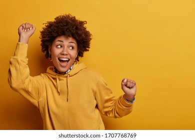 Photo of African American girl does lucky dance, raises hands up in hooray, feels like champion after getting triumph, gazes happily somewhere, has fun, feels rthythm of music, isolated on yellow wall